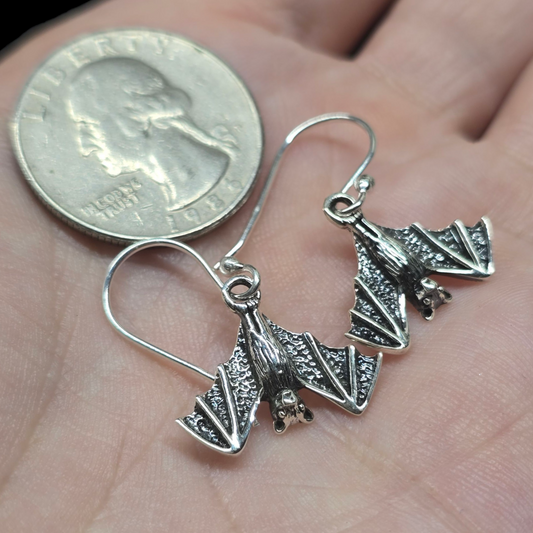 Silver bat-shaped earrings held between a hand and a quarter for size comparison.